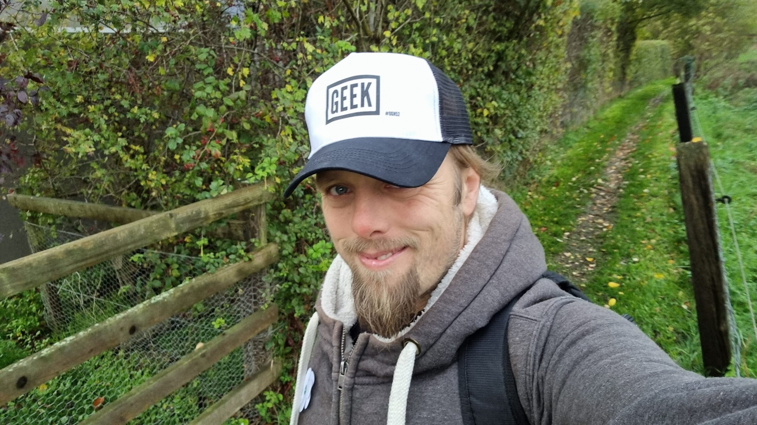 Full image of Dan, outdoors on a grassy path, wearing a grey hoodie. On his head is a "trucker cap" emblazoned with the word "GEEK" and, in smaller writing "#OGN52".