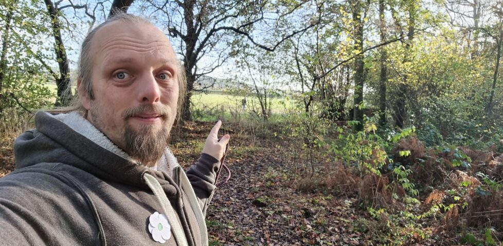 Dan, wearing a grey Three Rings hoodie with a white poppy pinned to it, holding the end of a dog lead, points out of the edge of the woodland he's in towards a humanoid figure in the distance.
