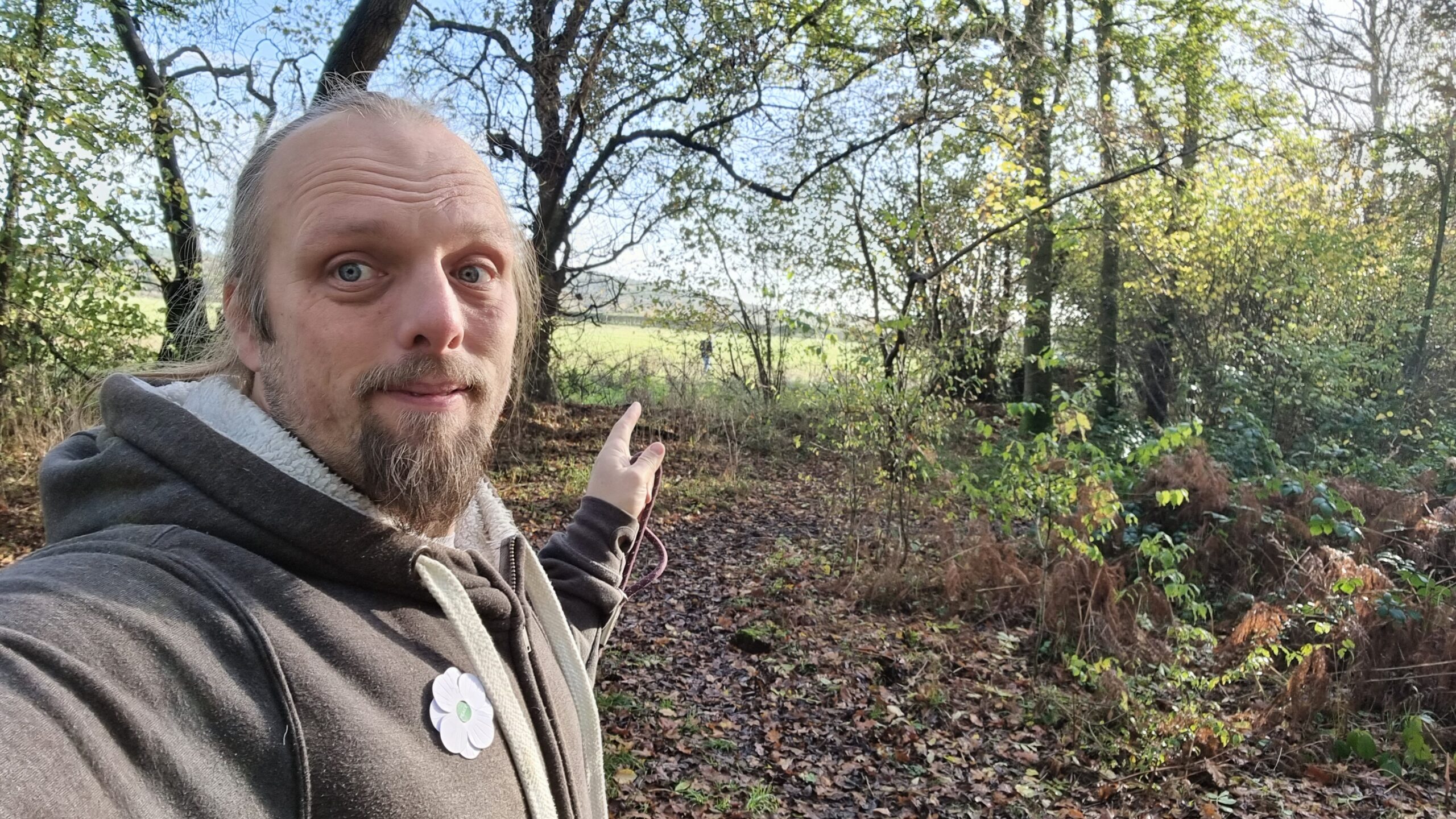 Full image of Dan, wearing a grey Three Rings hoodie with a white poppy pinned to it, holding the end of a dog lead, points out of the edge of the woodland he's in towards a humanoid figure in the distance.