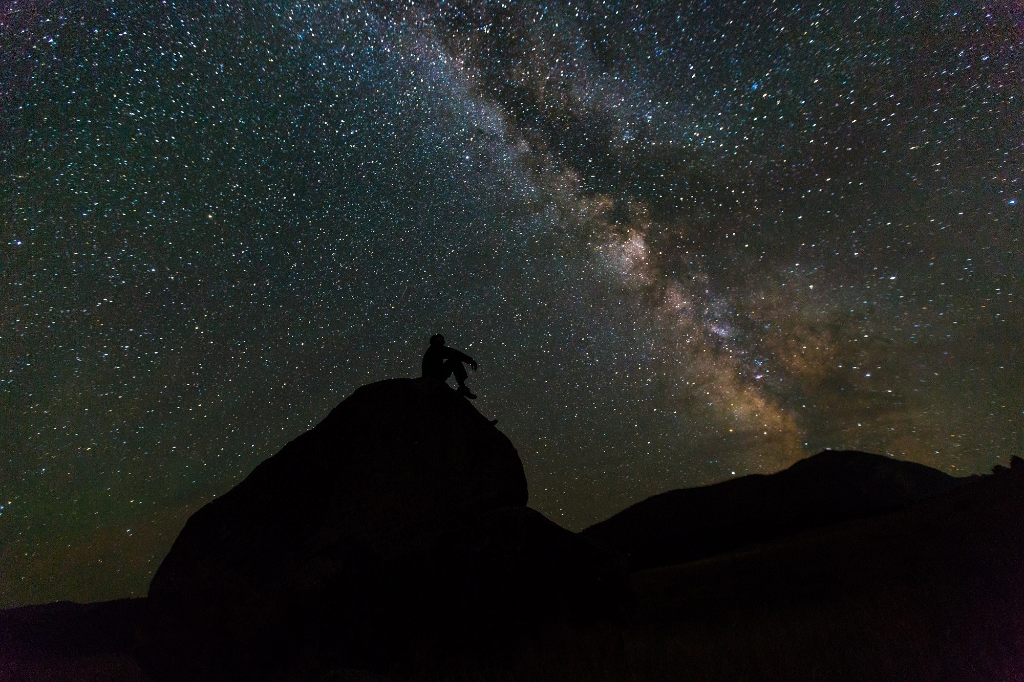 Full image of A silhouette of a person sits on a rock, gazing up at an incredible number of stars in an inky black sky.