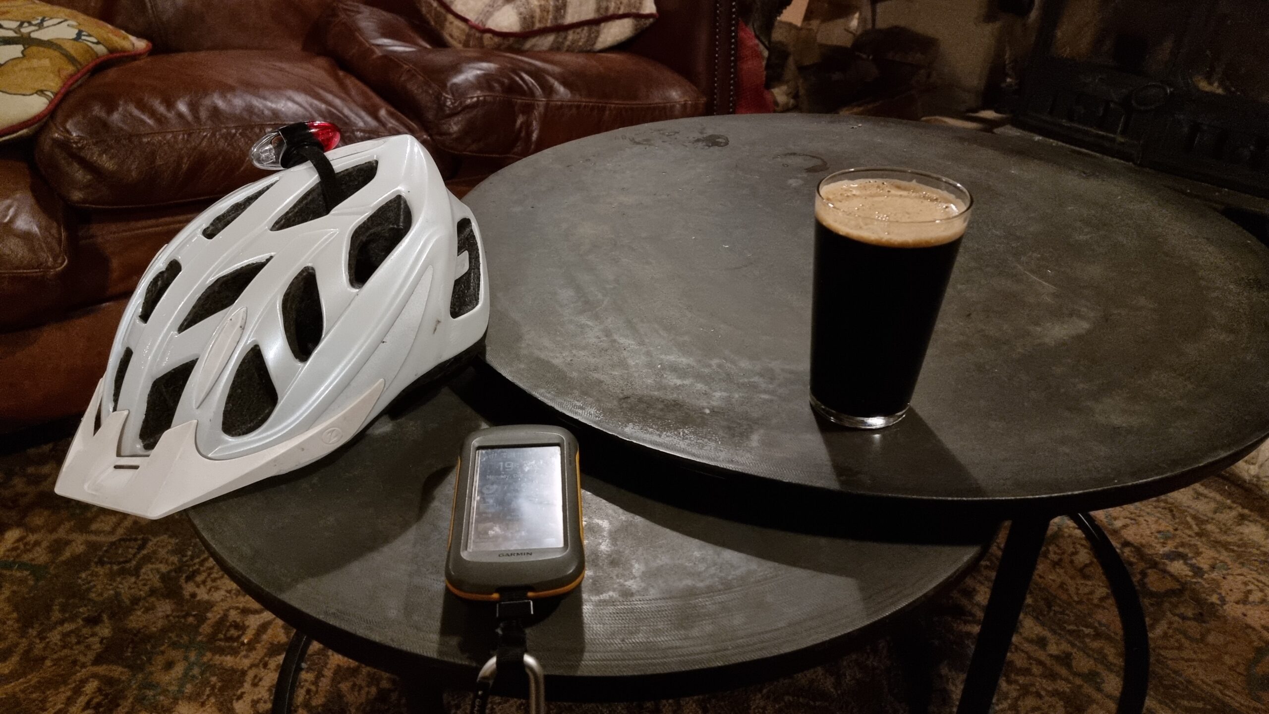 Full image of A round coffee table in front of a red leather sofa contains a cycle helmet, a GPS receiver (the first two digits of the time - 19 - are visible on its screen), and a pint of dark-coloured beer.