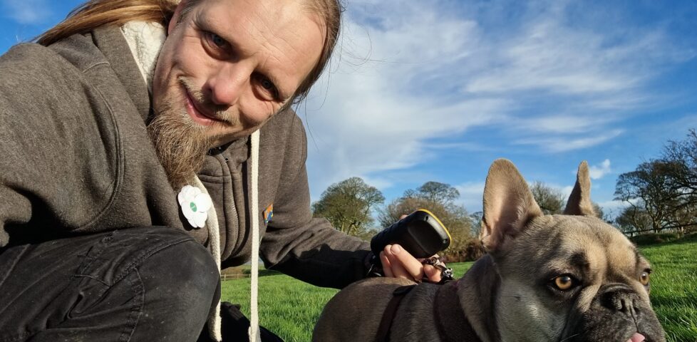 Dan - a man with a beard, wearing a grey fleece with a white poppy attached - crouches alongside his French Bulldog in a green field under a blue sky with a few wispy clouds.