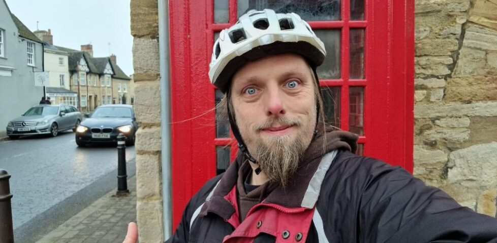 Dan, wearing a red-and-black jacket and a white cycle helmet, stands in front of a red telephone box on a village street.