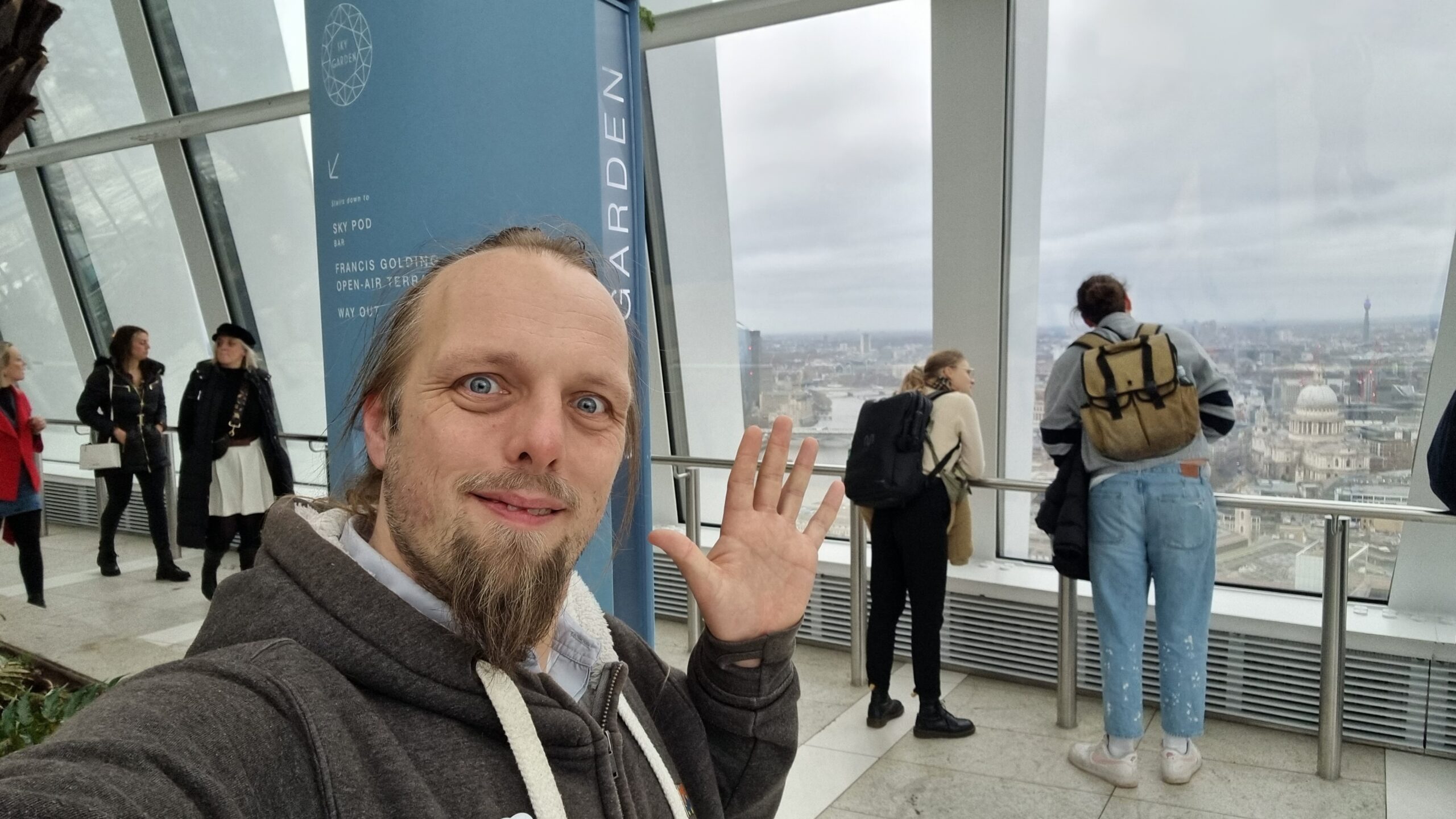 Full image of Dan waves from the Sky Garden; an aerial view of Central London is visible through the windows behind him.