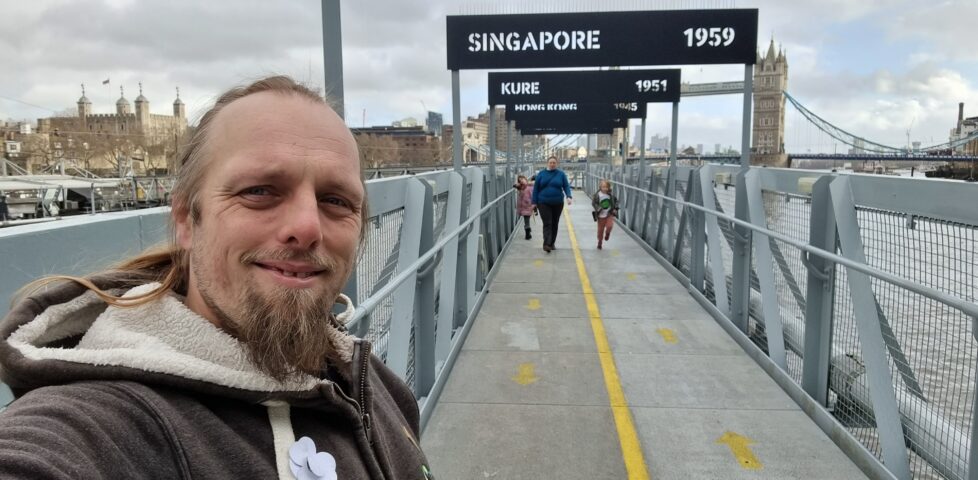 Dan stands on a pier leading out to the HMS Belfast, beneath a series of signs indicating where it served in particular years. In the distance, Ruth and the kids approach.