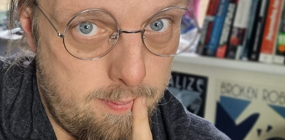 A white man wearing simple round spectacles with a silver wire rim, his finger to the corner of his mouth, looks into the camera.