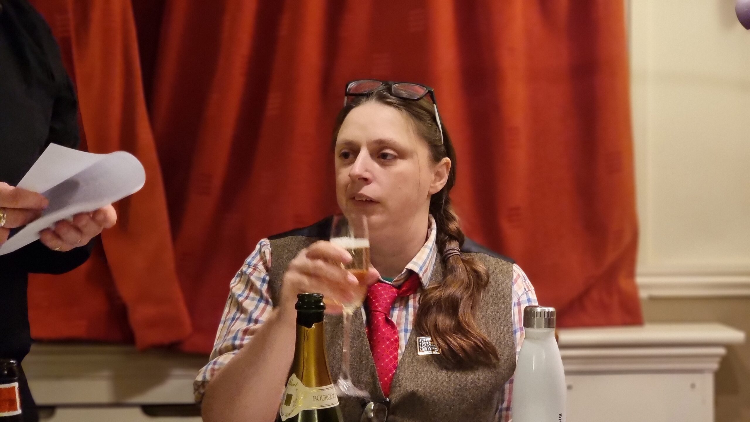 Full image of A woman dressed in a tweed waistcoat, glasses perched atop her head, sips a glass of champagne.