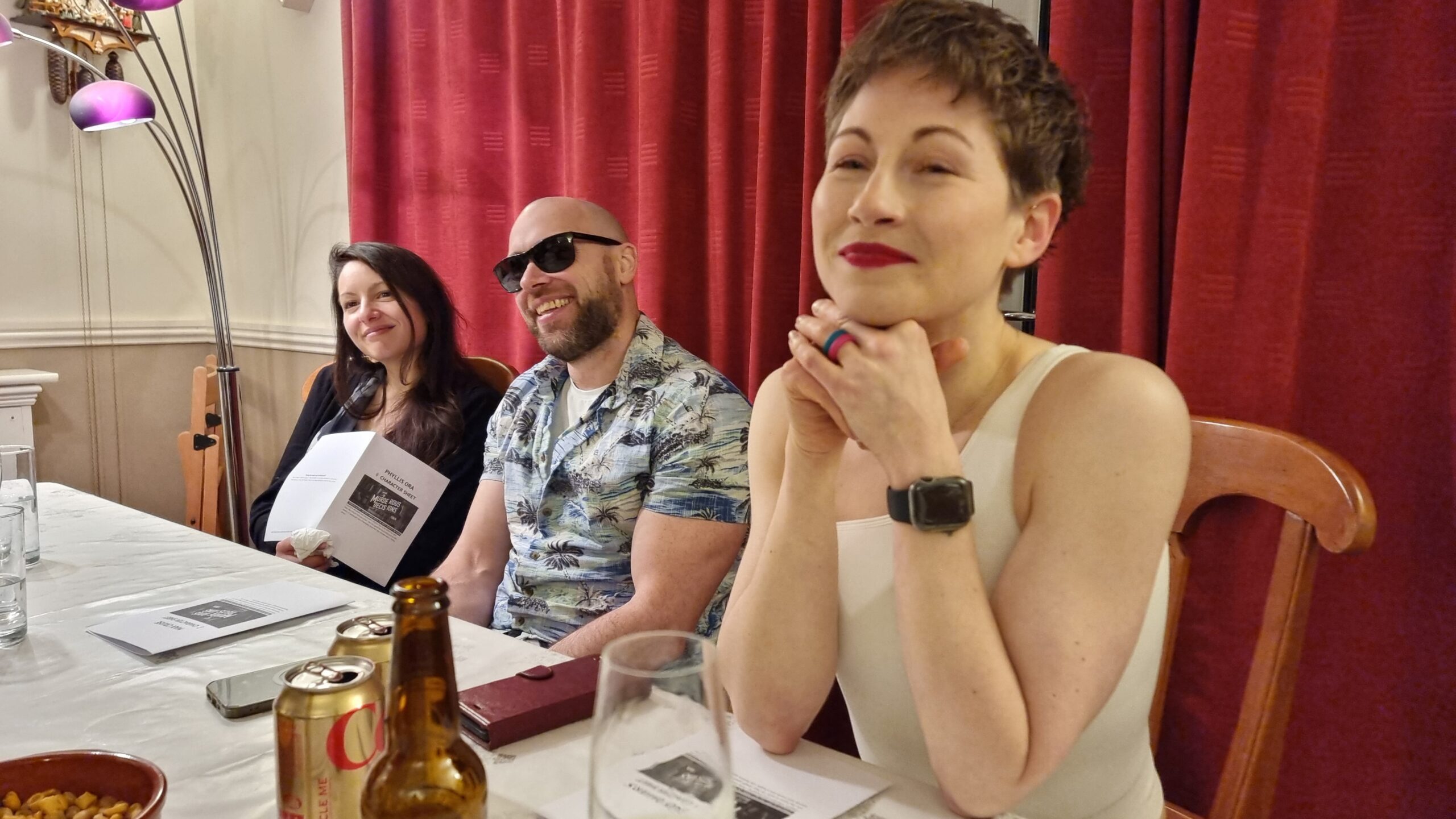Full image of Three people sit alongside a dining table: a woman with dark hair, holding her play notes, a man wearing sunglasses and a Hawaiian shirt, grinning, and a short-haired woman in a conforming white off-the-shoulder top and bright red lipstick. There are beer bottles, champagne flutes, and Coke cans on the table.