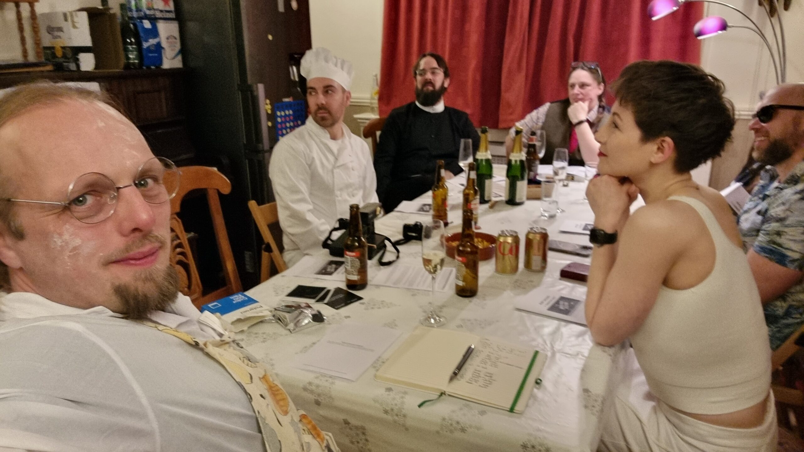 Full image of A group of people sit around a dining table; one man, wearing spectacles on his floury face, is taking the photo in "selfie" mode. The other visible characters appear to be a chef, a vicar, a librarian, a thrillseeker, and an aspiring starlet. With the exception of the photographer, they're all looking off to the side, listening to an unseen character speak.