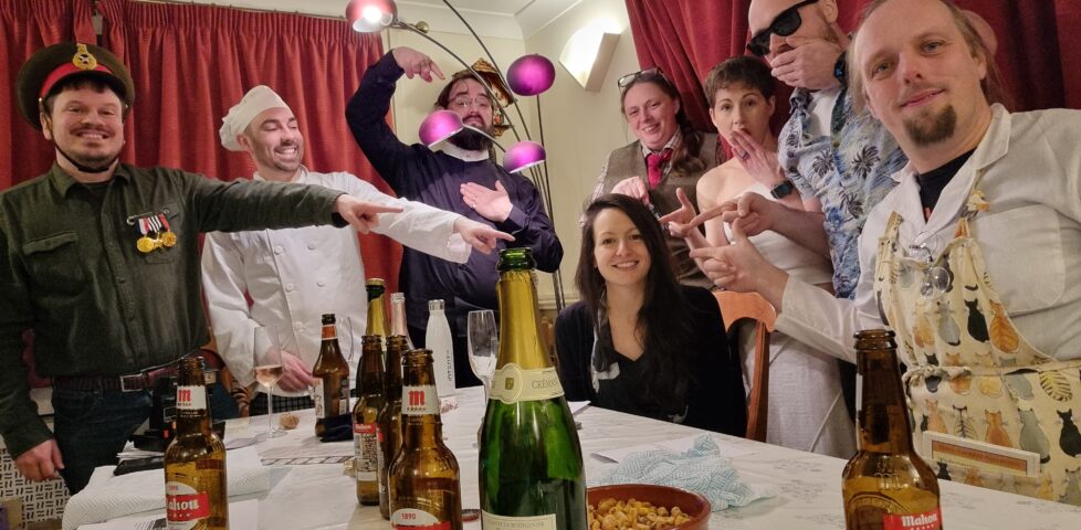 A party of unlikely characters stands around and points accusingly at the "murderer" among them: it's Kirsty.