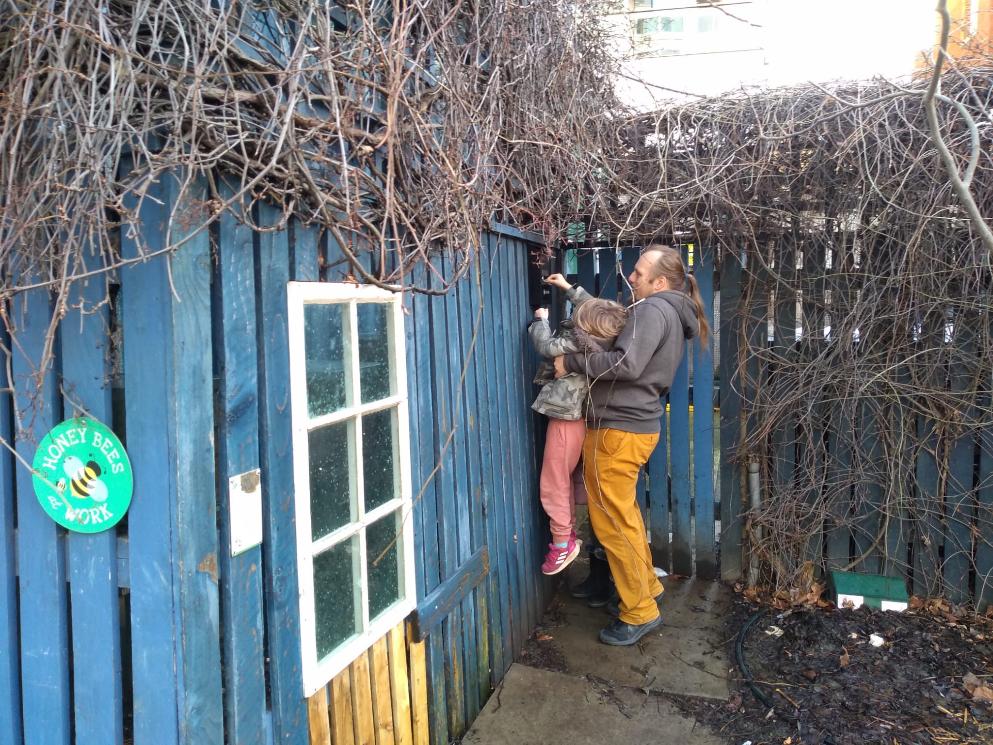 Full image of Alongside a blue fence, Dan lifts up a small boy so he can reach a wall-mounted geocache.