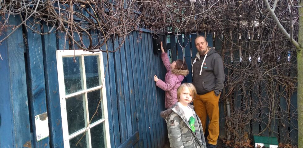 Dan stands with two children - a boy and a girl - near a high wall-mounted geocache; the girl is stretching to reach the cache container.