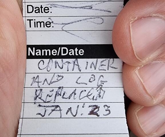 Fresh geocaching log book with a message stating that it's a replacement, added January 2023, held in a hand.
