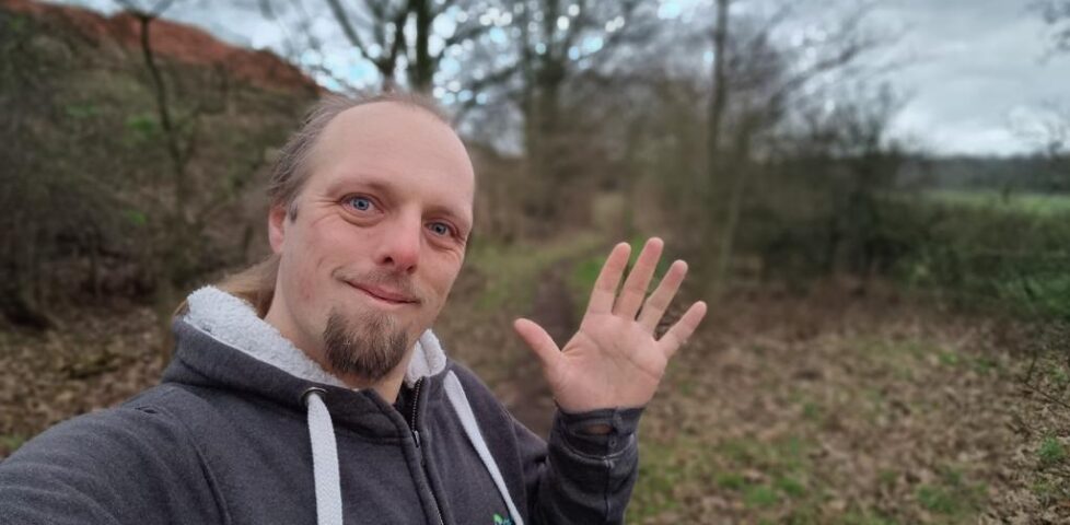 Dan, wearing a grey hoodie, waves to the camera from a dry wintery footpath.