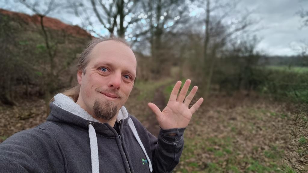 Dan, wearing a grey hoodie, waves to the camera from a dry wintery footpath.