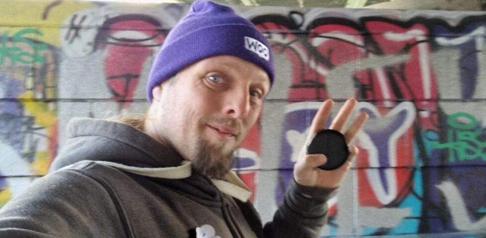Dan, wearing a grey hoodie and a purple "Woo" hat, holds a black puck-shaped geocache. Behind him, a concrete bridge support pillar is decorated with colourful graffifi.