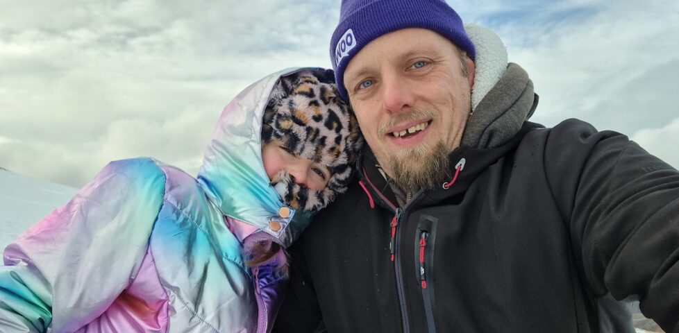 Dan, wearing a warm weatherproof black jacket and a purple "Woo" woolen hat, alongside a 9-year-old girl wrapped up in a faux-leapordskin hat and an iridescent coat, against a snowy hillside with rolling clouds.