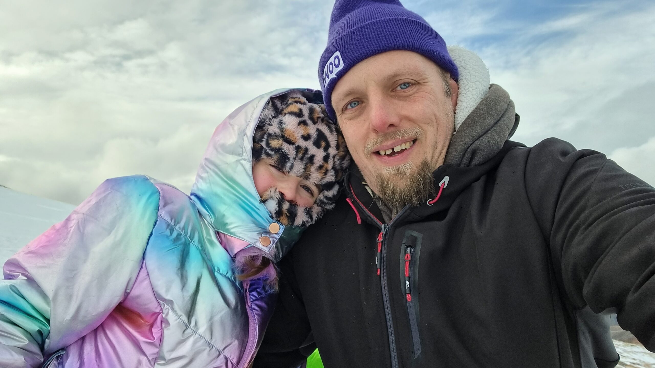 Full image of Dan, wearing a warm weatherproof black jacket and a purple "Woo" woolen hat, alongside a 9-year-old girl wrapped up in a faux-leapordskin hat and an iridescent coat, against a snowy hillside with rolling clouds.