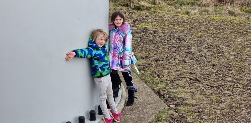 A smiling boy and girl in brightly-coloured coats hug the bottom of a very large wind turbine stem.