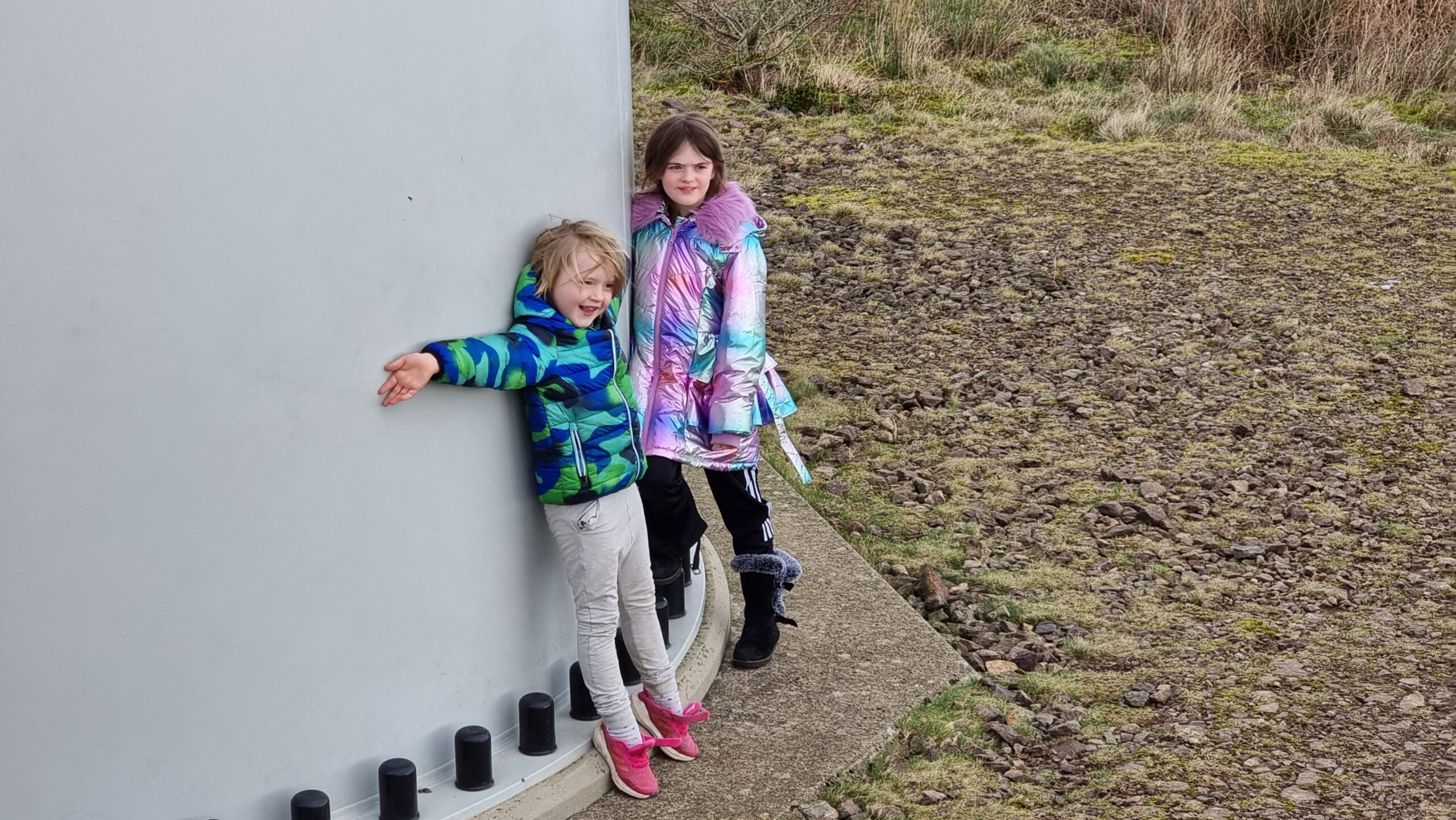 Full image of A smiling boy and girl in brightly-coloured coats hug the bottom of a very large wind turbine stem.