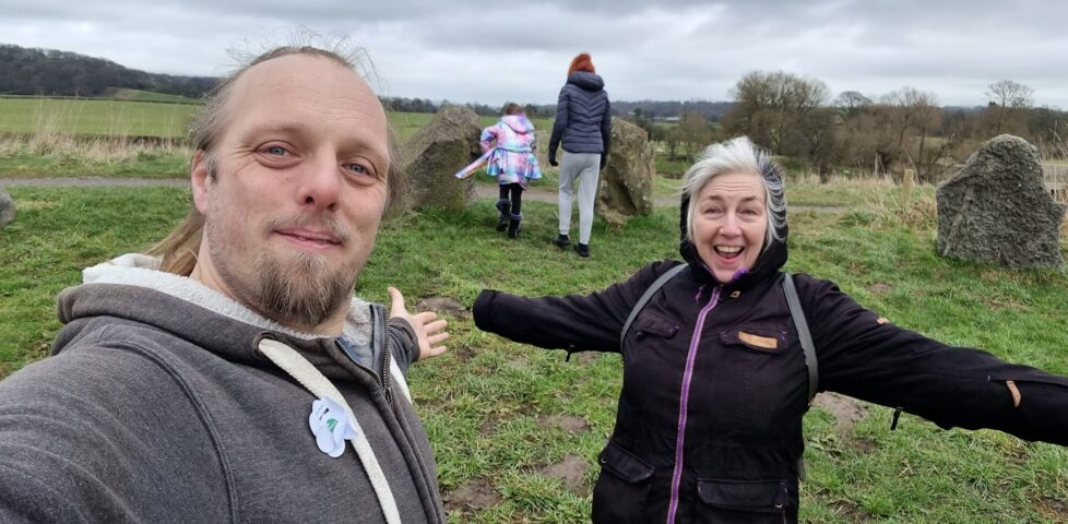 Dan and his mother stand in a stone circle, their arms spread wide and smiles on their faces.