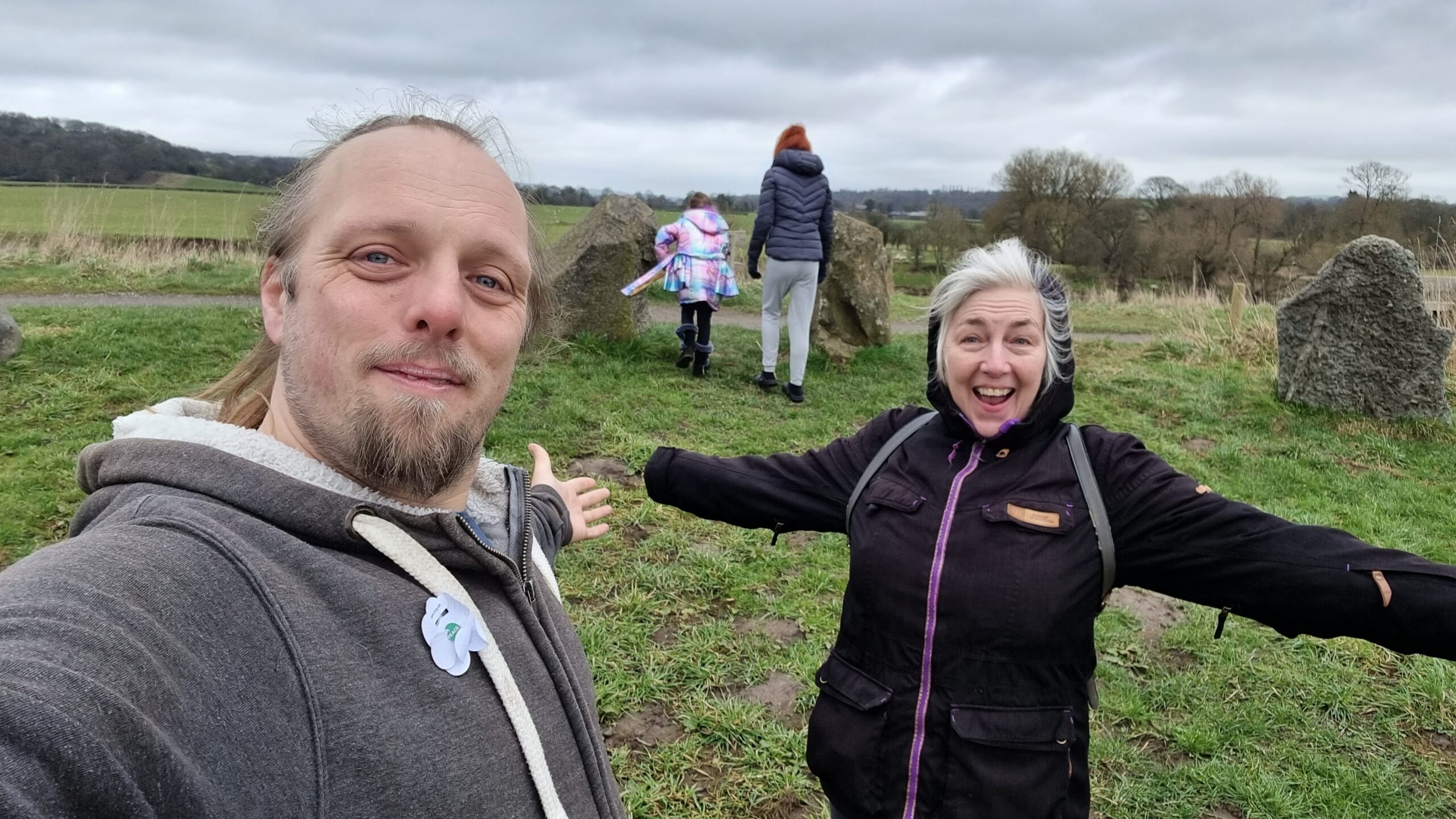 Full image of Dan and his mother stand in a stone circle, their arms spread wide and smiles on their faces.