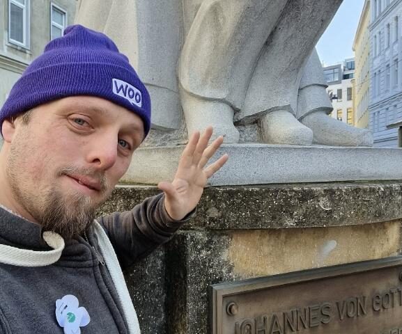 Dan, wearing a purple "Woo" wooly hat, stands in front of the base of a statue of Johannes von Gott.