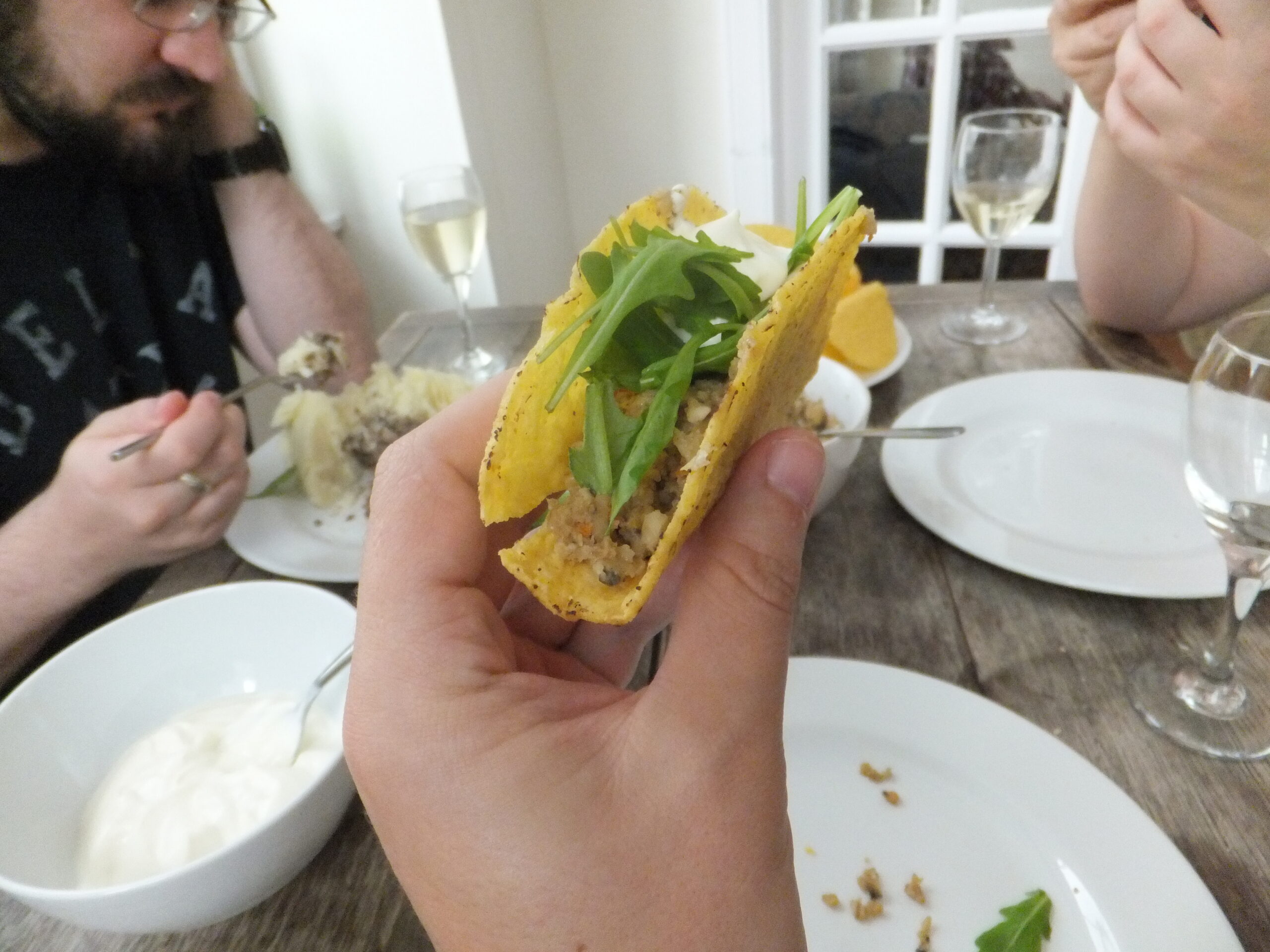Full image of A hand holds a crisp taco containing haggis, mashed potato, rocket, and a blob of sour cream. In the background, JTA can be seen eating his dinner in a more-conventional way: off a plate. There are glasses of wine on the table.