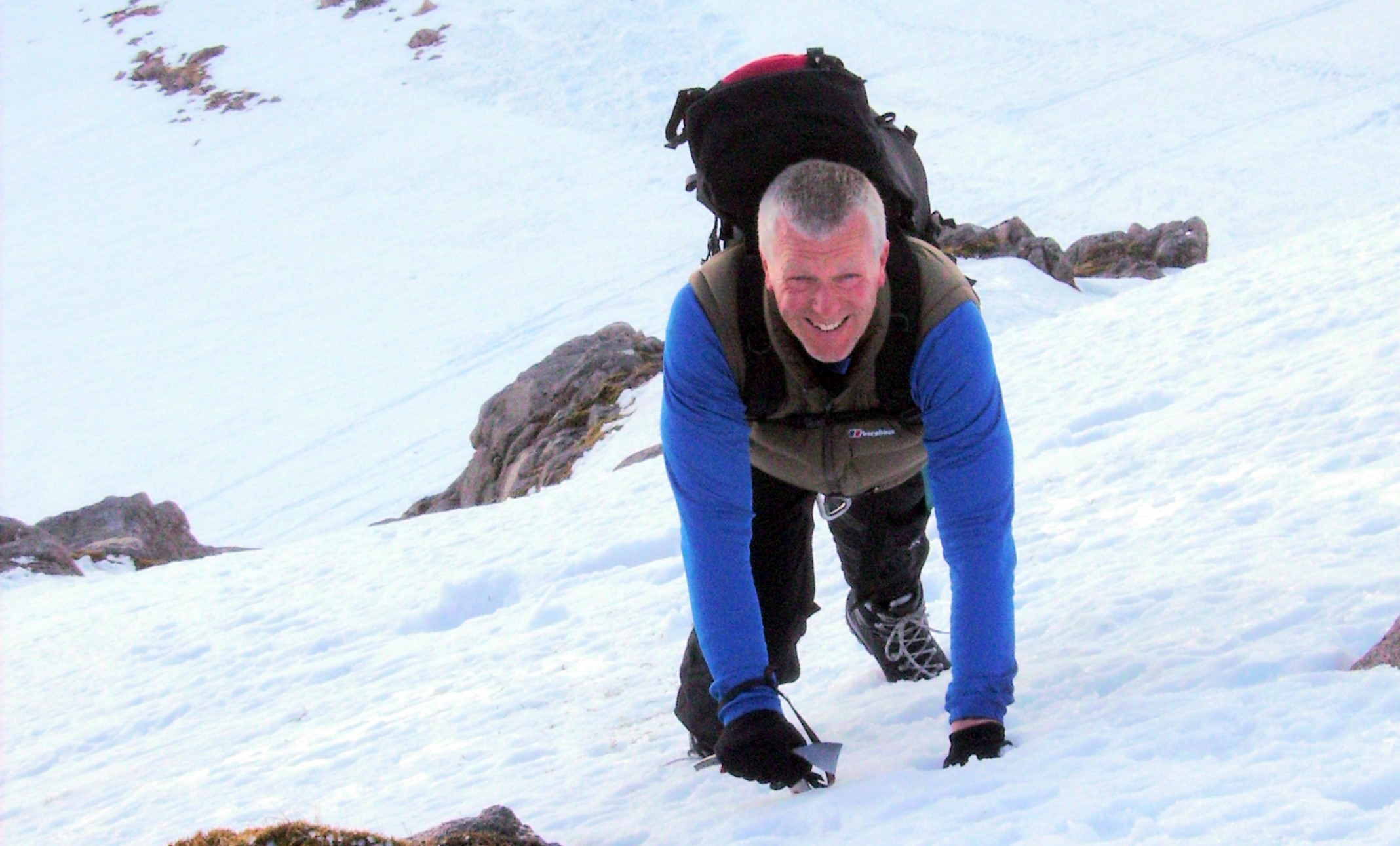 Full image of Seen from above, a man in his 50s wearing a large backpack uses mini ice axes to scramble up a steep hillside of powdered snow and rocks.