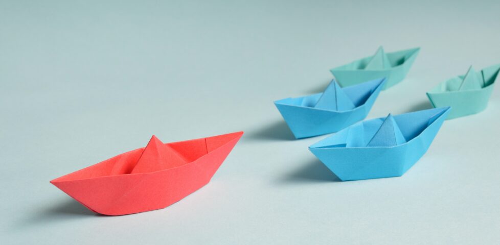 A red paper boat "sails" across a table, followed by four blue and green paper boats.