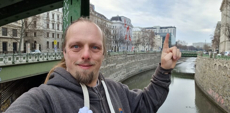 Dan stands on a bridge over the River Wein (before it meets the Danube), pointing to a window in the distance of a hotel.