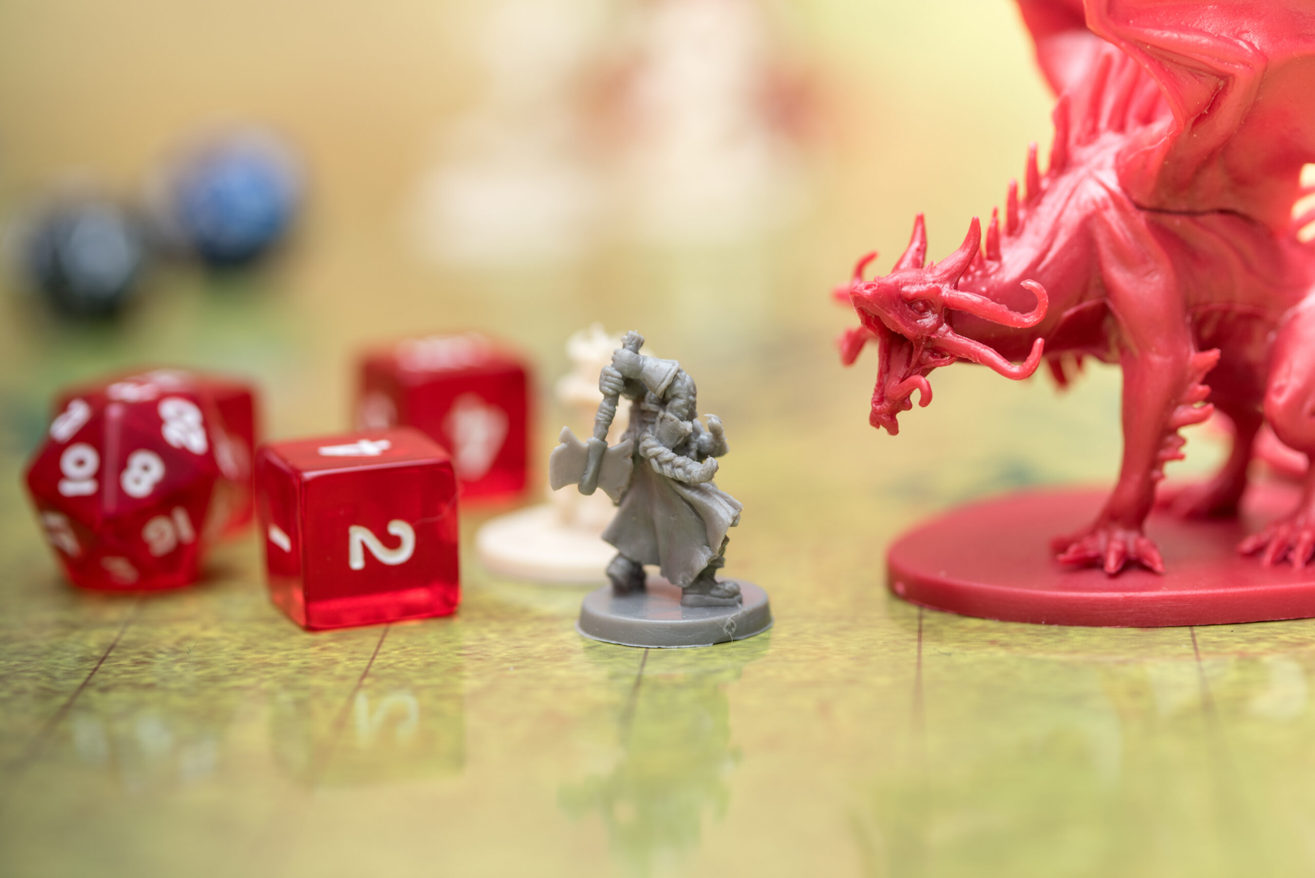 Full image of Photo with narrow depth of field, showing a tabletop roleplaying game. A grey plastic minature of a dwarf with a battle axe faces off against a large red minature of a dragon. Beside them, two glassy red six-sided dice and a similarly-coloured twenty-sided die can be seen.