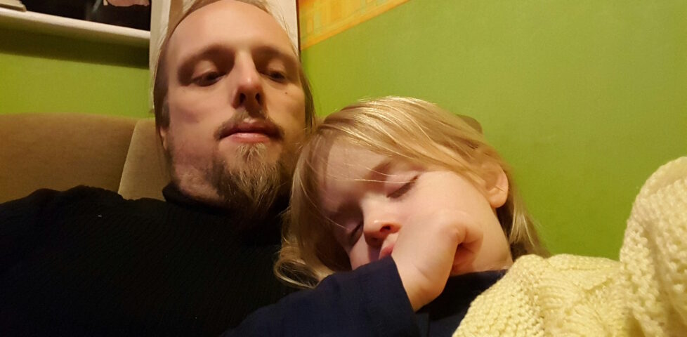 Dan, wearing a black jumper, sits on a green chair in a brightly-decorated bedroom. On his chest, a 2-year-old girl has fallen asleep, clutching a woolen yellow blanket and with her thumb in her mouth.