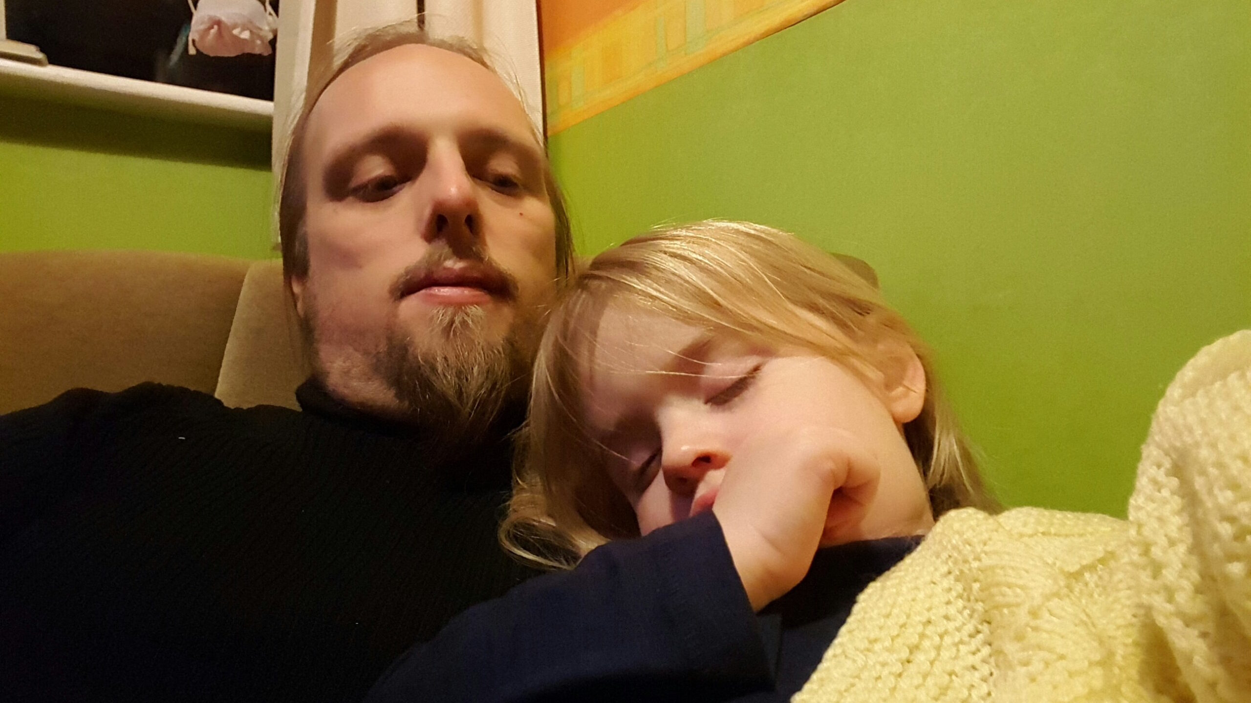 Full image of Dan, wearing a black jumper, sits on a green chair in a brightly-decorated bedroom. On his chest, a 2-year-old girl has fallen asleep, clutching a woolen yellow blanket and with her thumb in her mouth.