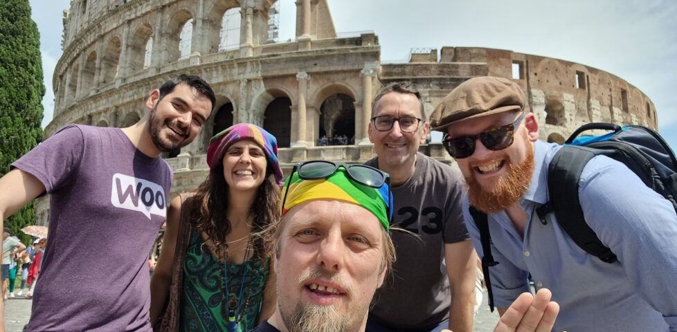 In front of the Colosseum in Rome, Dan - wearing a rainbow-striped bandana atop which his sunglasses are perched - takes a selfie. Behind him stand a man with dark hair and a closely-trimmed beard wearing a purple "woo" t-shirt, a woman with long brown hair wearing beads and a multicoloured dress, a man wearing spectacles and a dark t-shirt on which the number "23" can be made out, and a man in sunglasses with a ginger beard, wearing an open blue shirt.