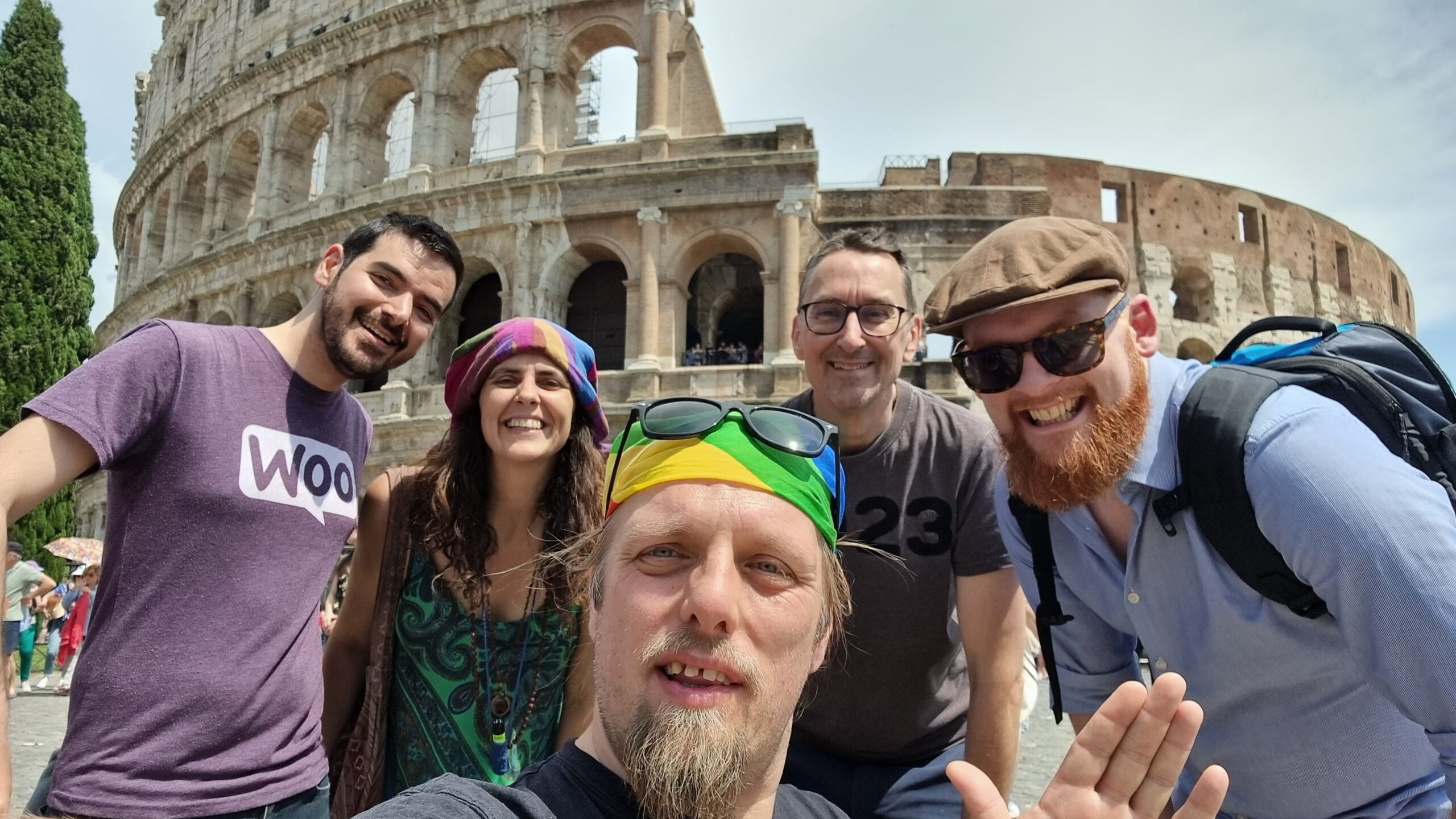 Full image of In front of the Colosseum in Rome, Dan - wearing a rainbow-striped bandana atop which his sunglasses are perched - takes a selfie. Behind him stand a man with dark hair and a closely-trimmed beard wearing a purple "woo" t-shirt, a woman with long brown hair wearing beads and a multicoloured dress, a man wearing spectacles and a dark t-shirt on which the number "23" can be made out, and a man in sunglasses with a ginger beard, wearing an open blue shirt.