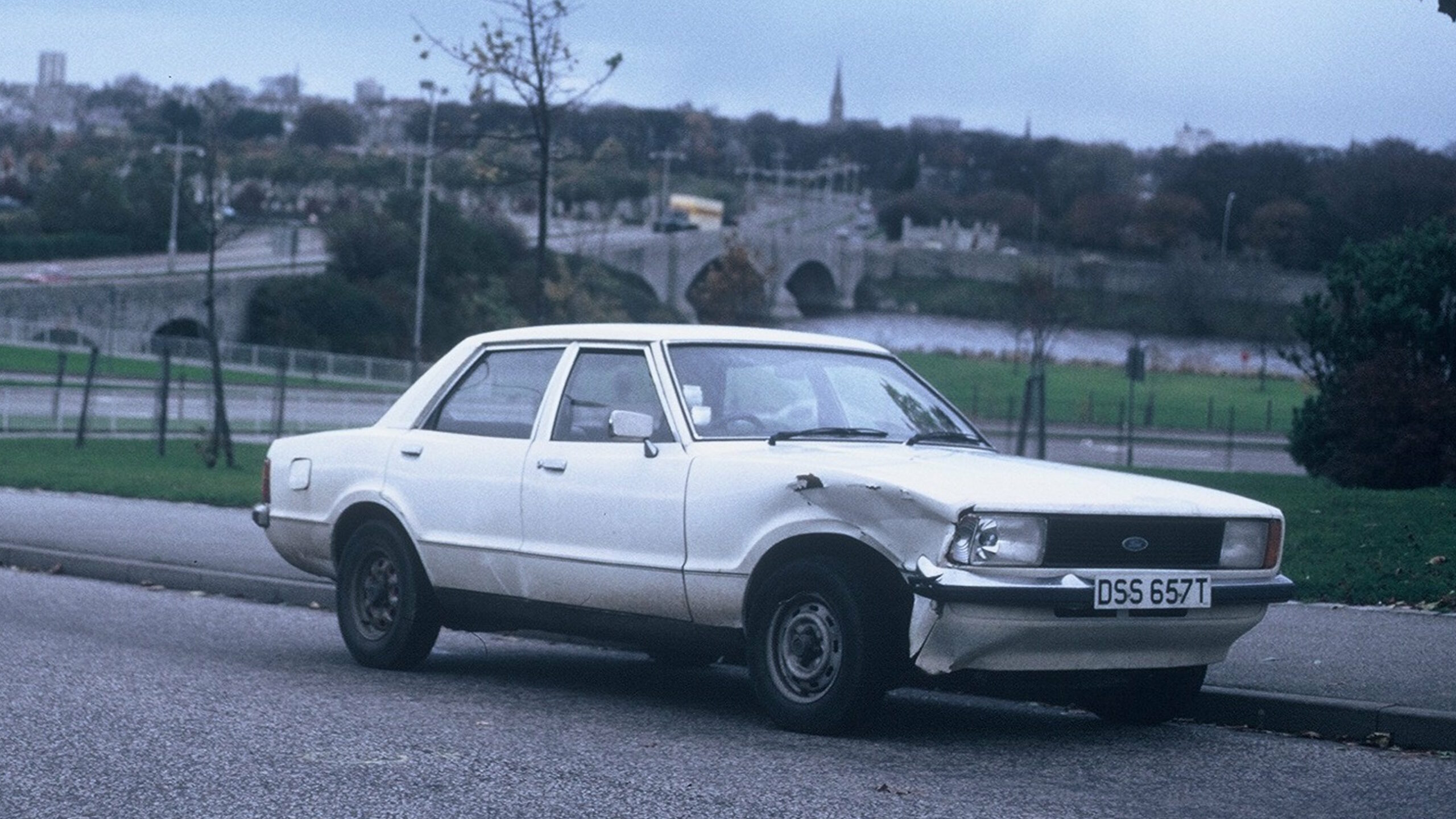 Full image of A boxy 1979 white Ford car, number plate DSS 657T with a badly dented and somewhat corroded front wheel arch on the drivers' side, sits empty and parked at the side of an otherwise empty asphalt strreet. In the background, under grey skies, a city skyline can be made out with houses, tower blocks, and a church steeple, on the other side of an arched river bridge. The leaves are early-autumn coloured: mostly greem, but with some brown appearing and a handful of bare branches exposed.