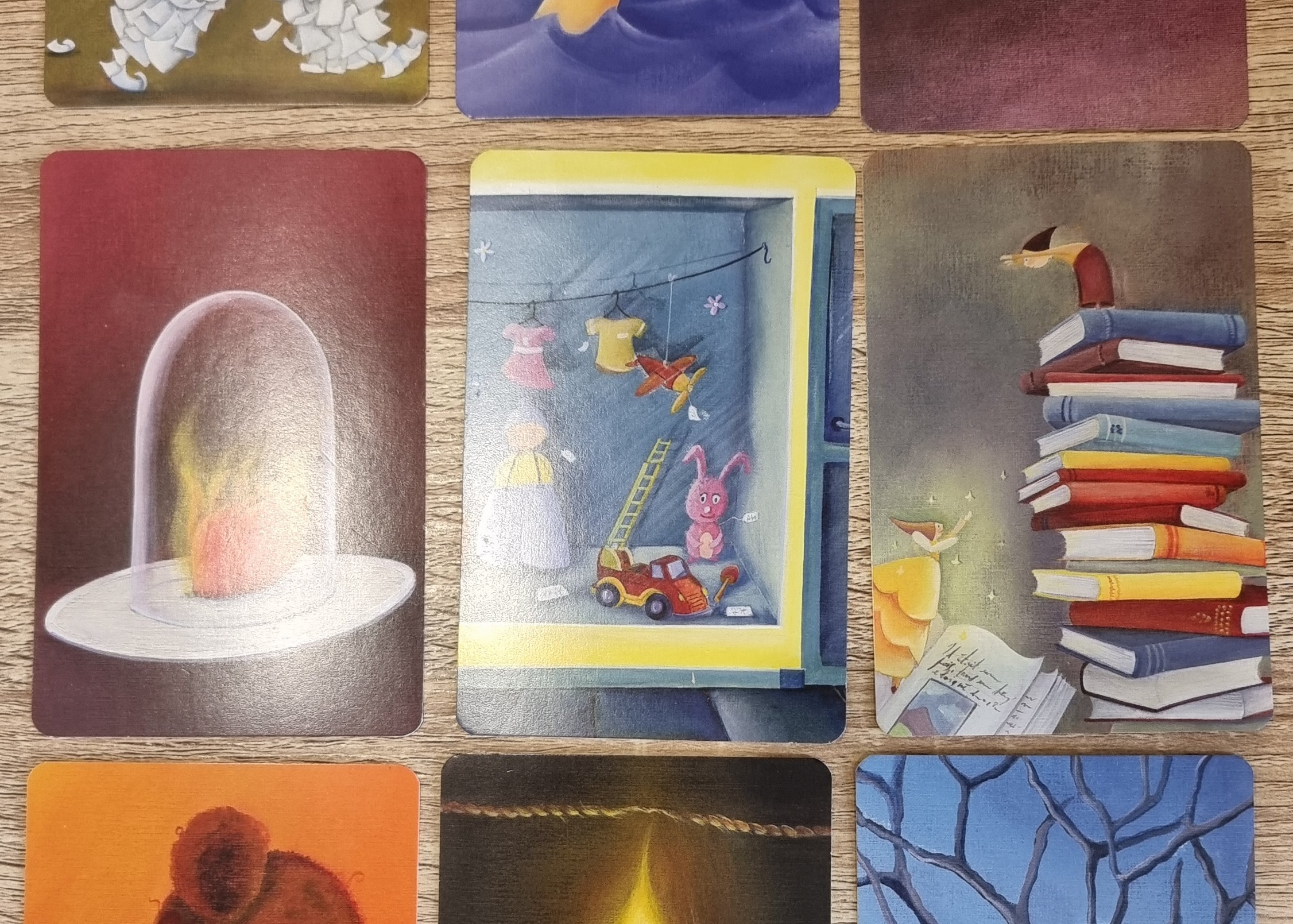 Full image of A trio of Dixit cards within a grid of nine. From left to right, they show: a heart, on fire, beneath a glass jar; a cubbyhole containing childrens' toys; a fairy leaping from a book towards a small person atop a stack of books.