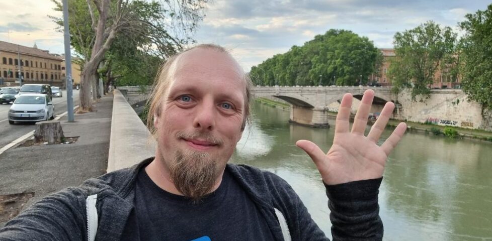 Alongside the River Tiber, with a wide-arched stone bridge in the background, Dan sits on a wall and waves to the camera.
