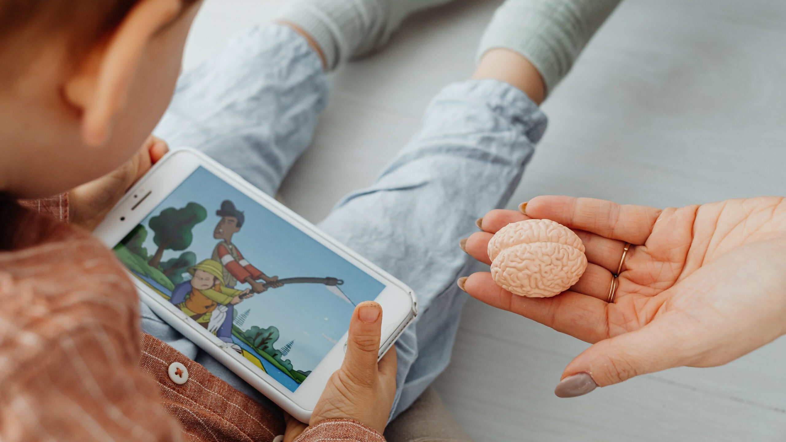 Full image of A small child, sitting on the floor, uses a mobile phone to watch a cartoon of two people struggling to pull a fishing rod. A feminine hand with brown-painted nails and rings on two fingers reaches in to offer the child a minature model of a human brain.