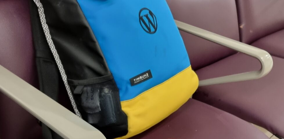 A modest-sized backpack in blue and yellow, with a WordPress logo stiched on, sits on an airport departure lounge bench. Alongside it is a burgundy-coloured British passport.