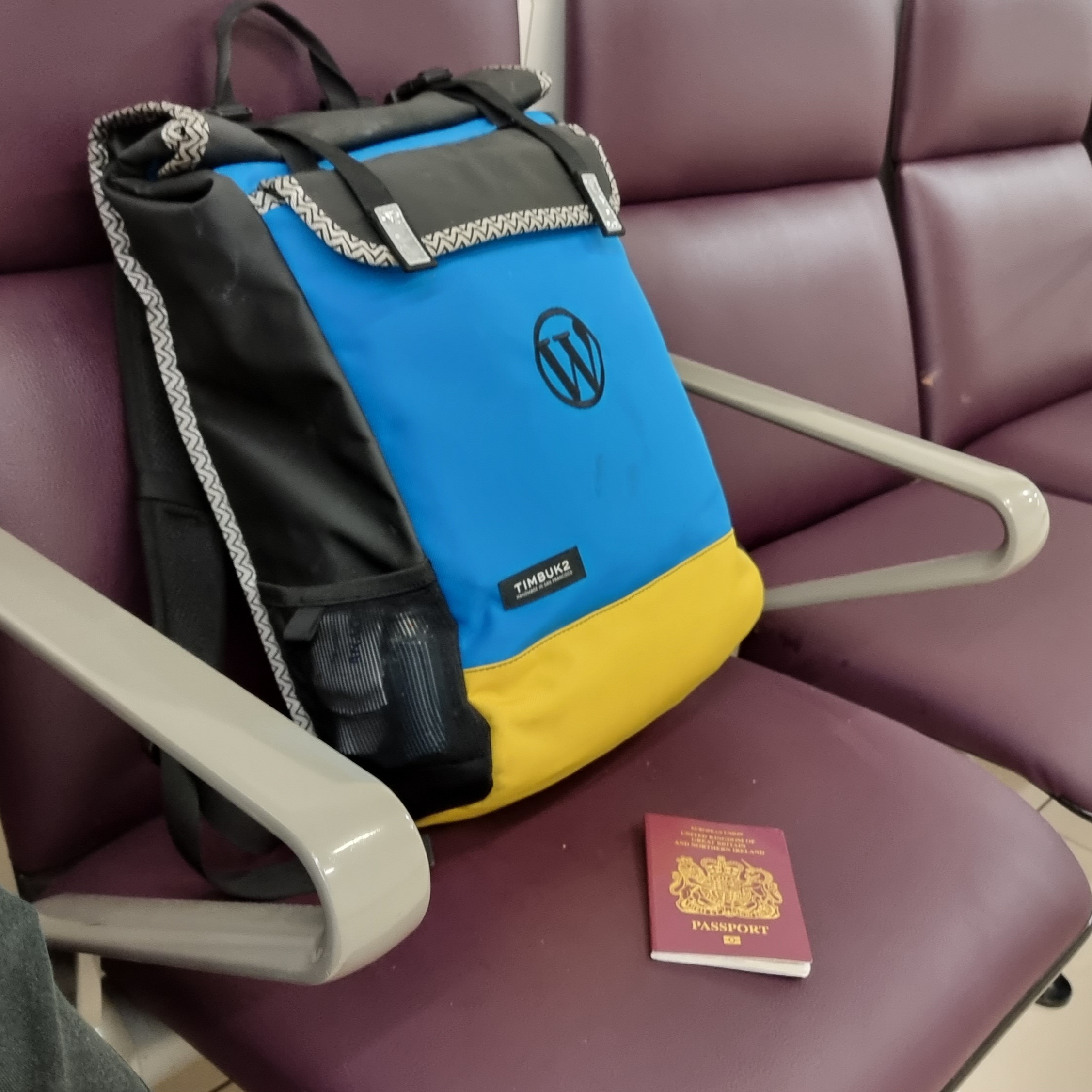 Full image of A modest-sized backpack in blue and yellow, with a WordPress logo stiched on, sits on an airport departure lounge bench. Alongside it is a burgundy-coloured British passport.