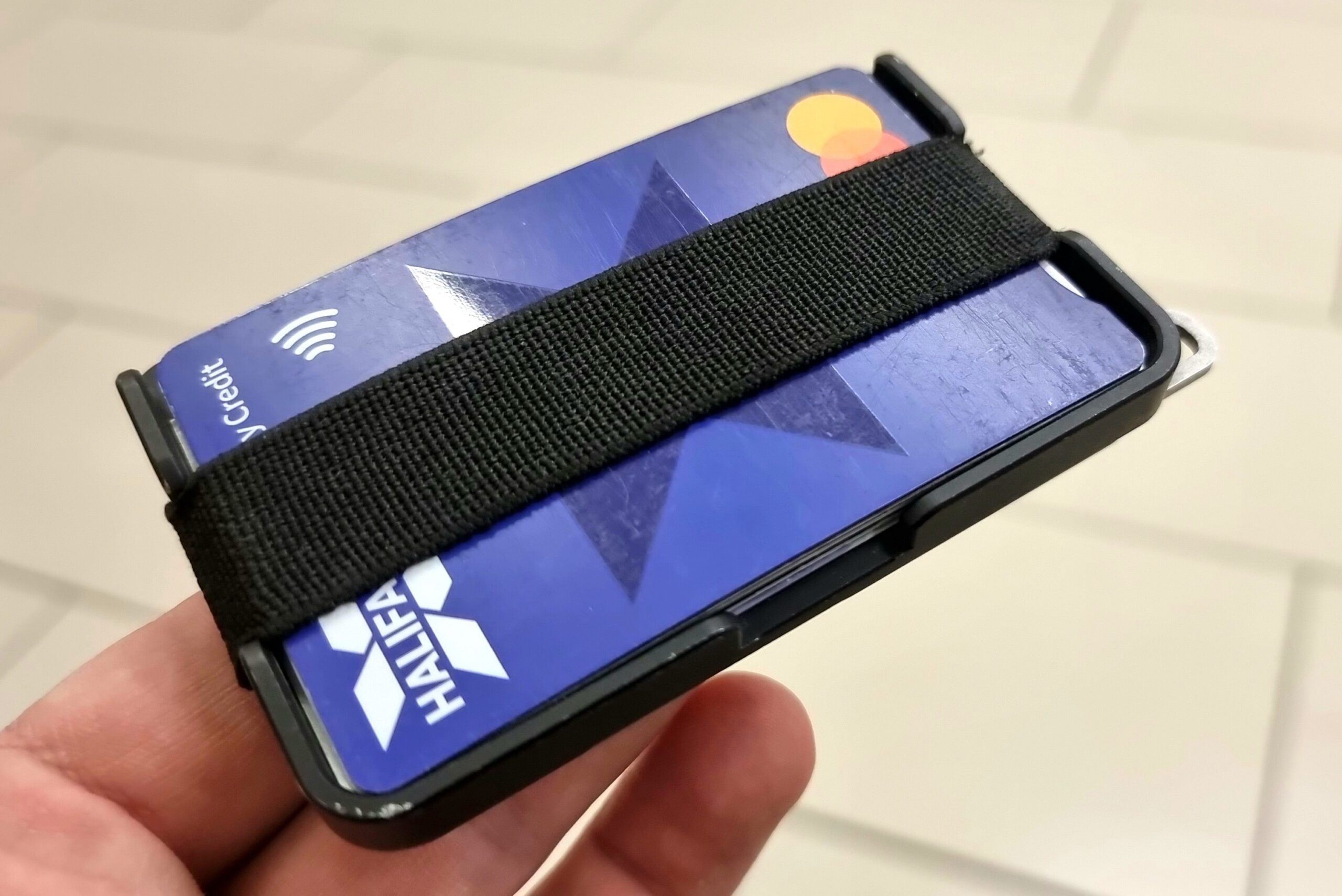 Full image of Minimalist carbon fibre wallet, balanced on two fingertips, with parts of a Halifax Mastercard credit card showing from behind an elasticated band.