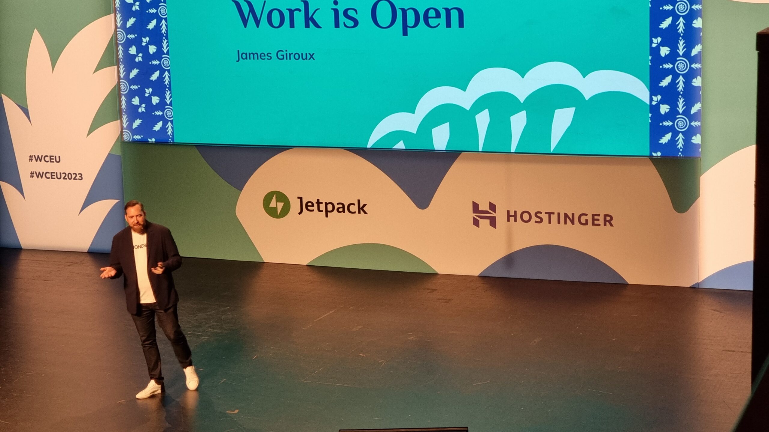 Full image of A man in a white t-shirt and dark jacket stands on a stage in front of a screen; the bottom line of the words on the screen can be seen to read "Work is Open".