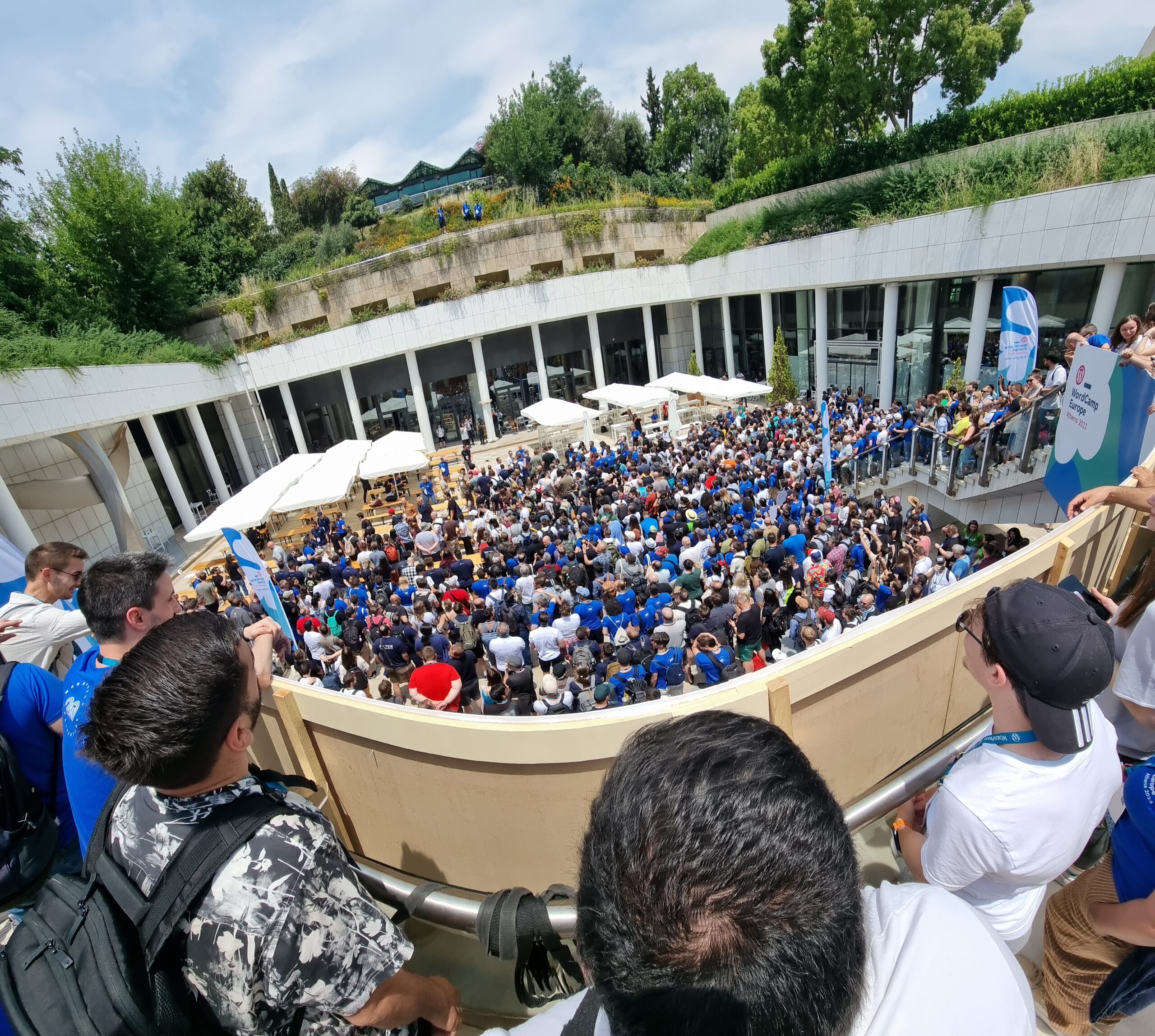Full image of An enormous crowd shuffles tightly into a courtyard. A trio of blue-shirted photographers stands atop a building opposite them.