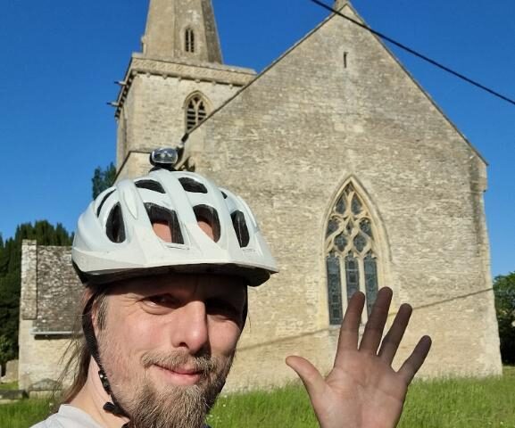 Dan, wearing a cycle helmet, stands in front of a church steeple under a bright blue sky, waving.
