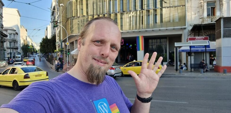 Dan, wearing a purple shirt with a WordPress logo and a pride rainbow, waves to the camera in front of a hotel with pride rainbow banners hanging from its pillars.