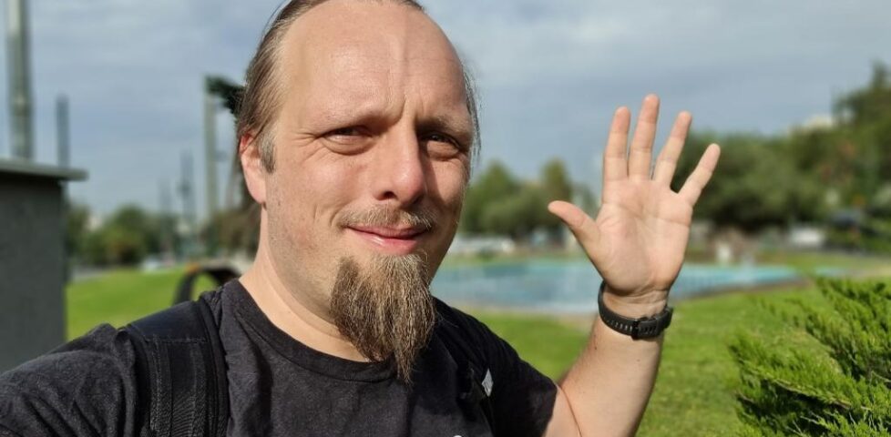 Dan, in a green park with water fountains, waves at the camera.