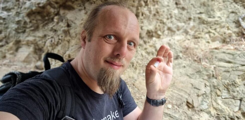 Dan, wearing a black t-shirt and a backpack, holds a tiny plastic container with a red lid. Behind him is a rough rock wall.