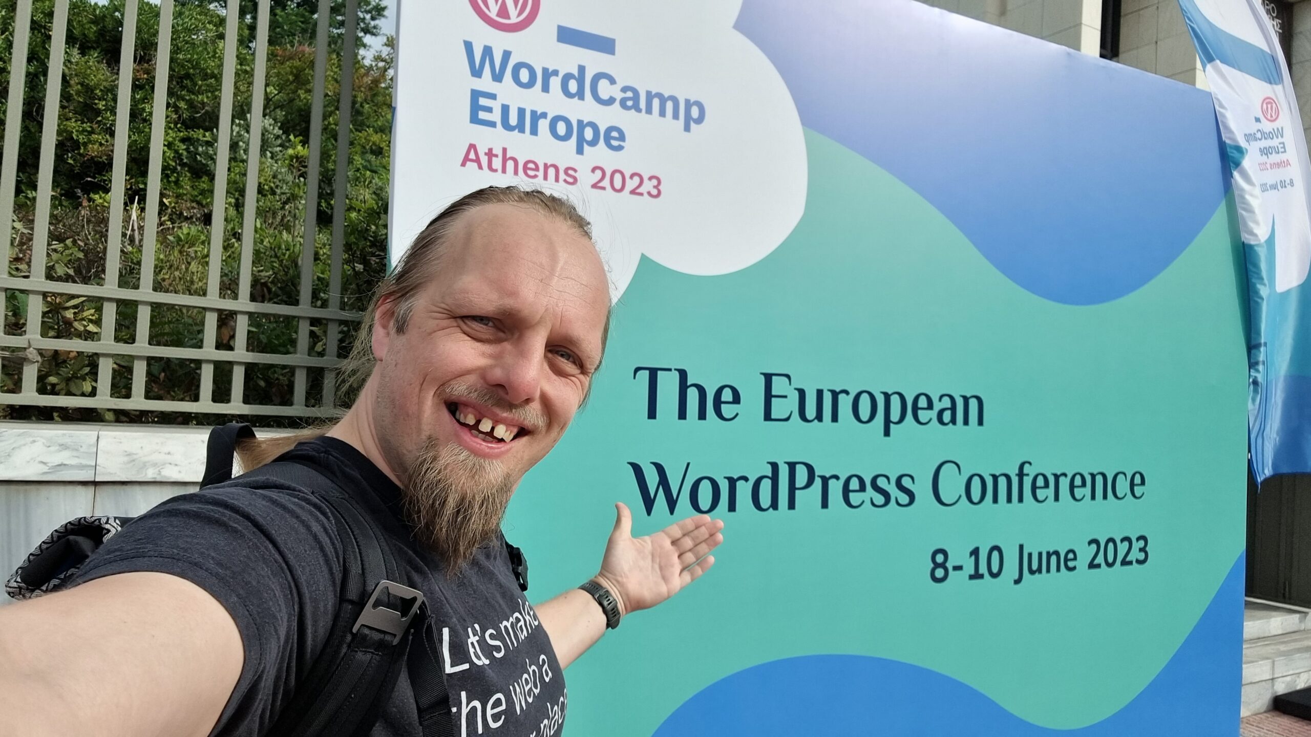 Full image of Dan, wearing an Automattic "Let's make the Web a better place" t-shirt, stands in front of a banner welcoming attendees to WordCamp Europe Athens 2023.